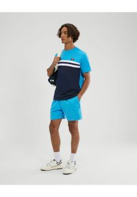 T-shirt color block blu e blu navy con strisce bianche; abbinato a pantaloni corti blu coordinati; il modello tiene una borsa; sneakers bianche con accenti neri.