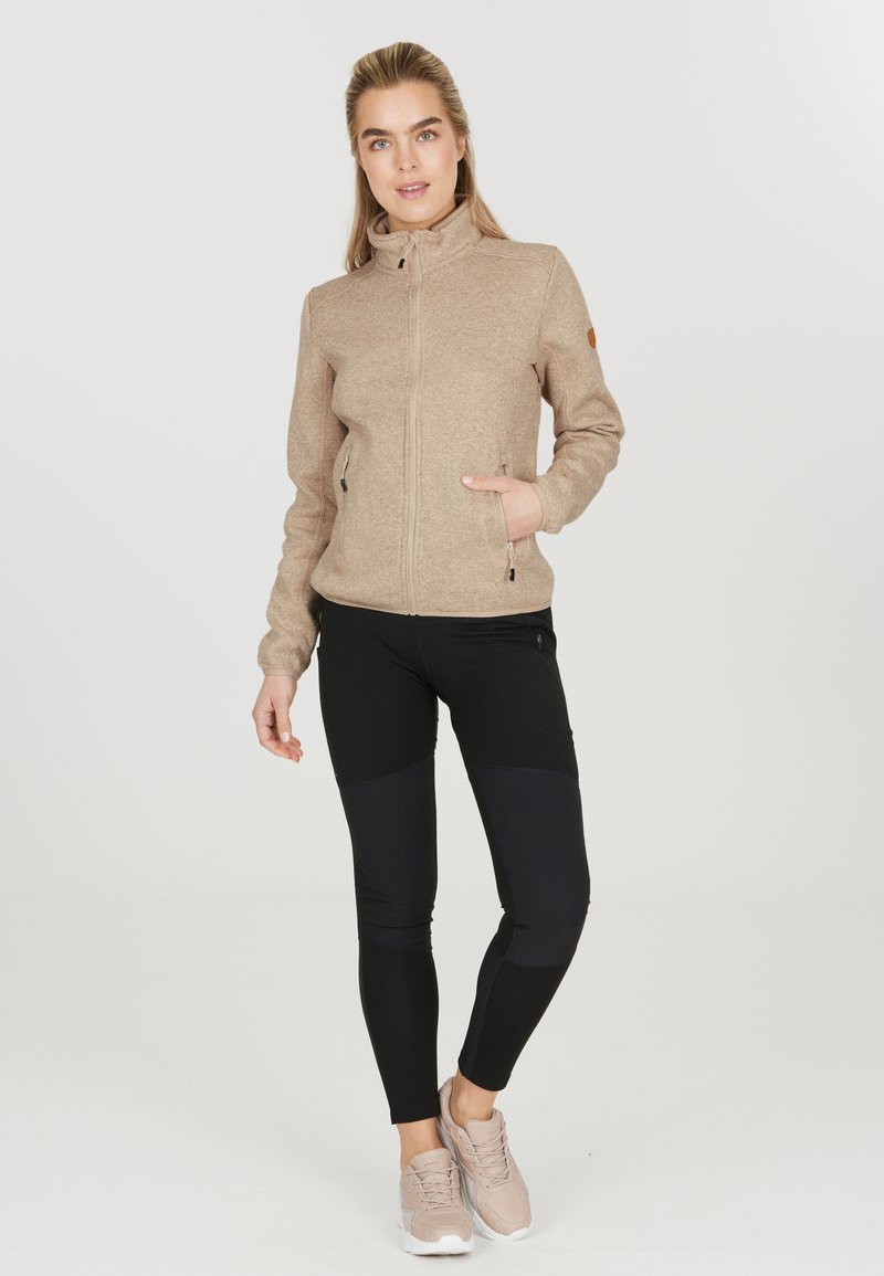 Veste beige zippée avec un col haut, deux poches latérales, et leggings noirs avec des accents texturés. Chaussures : sneakers beige clair.