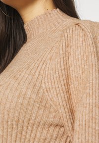 Pull en maille côtelée beige avec un col montant et des manches légèrement bouffantes, mettant en valeur une texture douce et une silhouette ajustée.