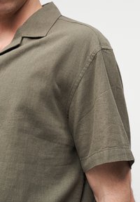 Άνδρας που φοράει πουκάμισο από λινό ελιάς με κοντό μανίκι και γιακά τύπου camp, καδριασμένος ώστε να φαίνεται το πάνω μέρος του στήθους και το δεξί χέρι σε απλό φόντο.