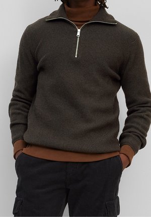 Mężczyzna w ciemnooliwkowym prążkowanym swetrze z półzamkiem, nałożonym na brązowy golf, noszący czarne sztruksowe spodnie typu cargo, z rękami w kieszeniach, na jasnym tle.