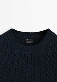 Pull en coton bleu marine à mailles torsadées avec col rond côtelé, montrant les étiquettes de la marque et du matériau à l'intérieur du col.