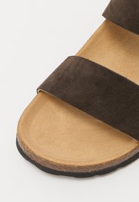 Brune semskede sandal med to brede remmer, en flad tannfarget såle og en svart gummisåle, som fremhever et enkelt, allsidig design.