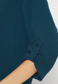 Camicia teal con bottoni e maniche arrotolate. Realizzata in un tessuto testurizzato che mostra una leggiera lucentezza. Presenta un solo bottone sulla manica.