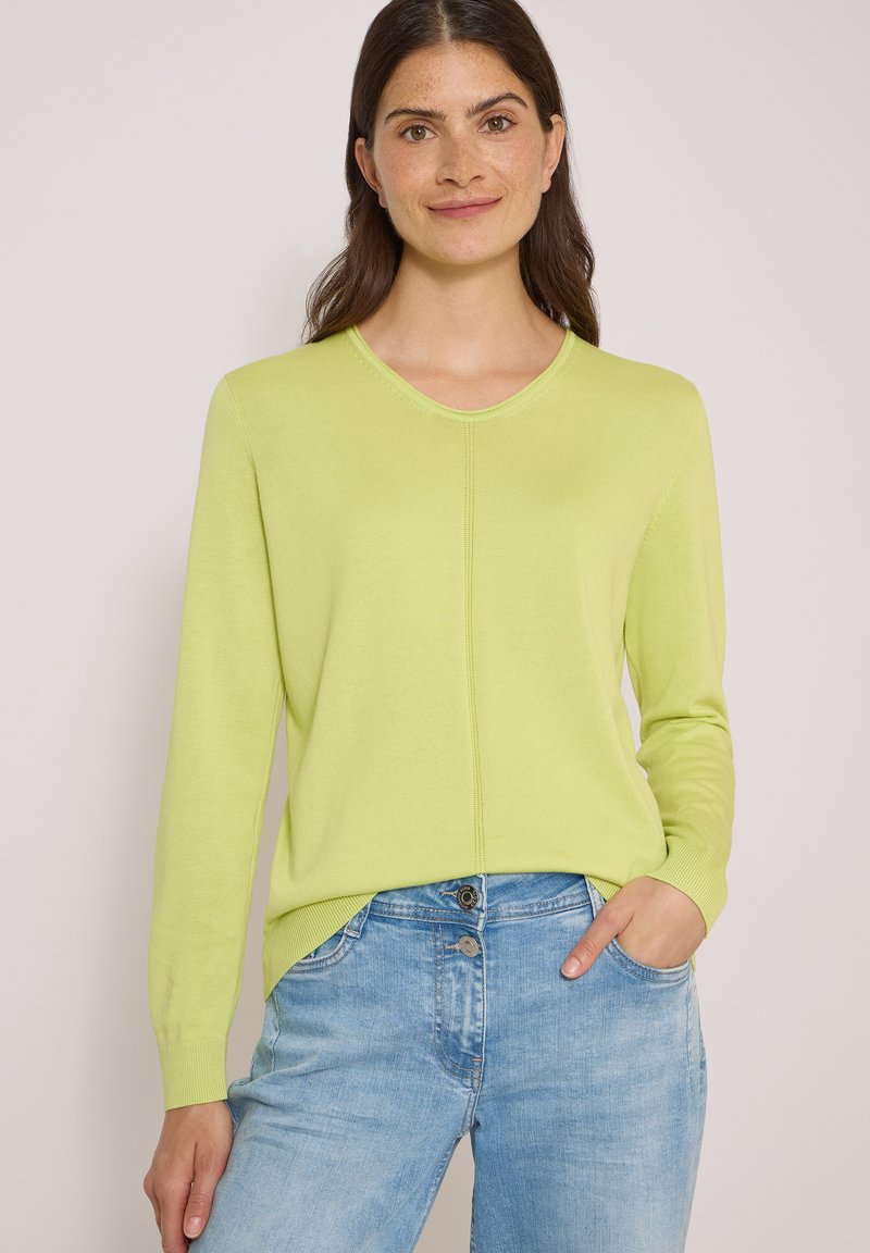 Femme aux longs cheveux bruns portant un pull vert clair à manches longues et un jean bleu clair, debout avec une main dans la poche devant un fond uni.