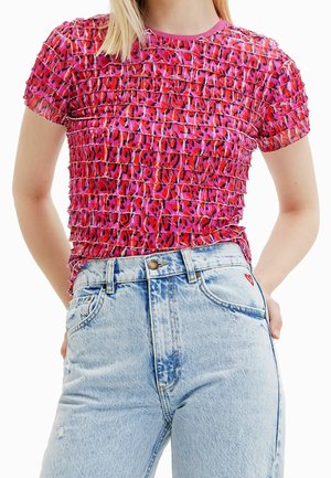 Kobieta nosząca różowo-czerwony krótki top z falbanami z abstrakcyjnym nadrukiem oraz jasno niebieskie jeansy z wysokim stanem z małym czerwonym serduszkiem.