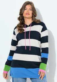 Vrouw met lang golvend haar, gekleed in een zwart-wit-blauw-groen gestreepte hoodie met roze trekkoorden en lichte blauwe jeans, staand tegen een witte muur.