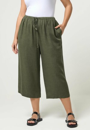 Frau trägt olivgrüne, weit geschnittene, knöchellange Hose mit Kordelzugbund und schwarzen Sandalen mit Schnalle, steht mit der Hand in der Tasche.