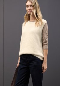 Un pull en tricot pelucheux de couleur crème clair superposé sur une chemise beige à manches longues, associé à un jean bleu foncé ; sans motifs ni éléments métalliques notables.