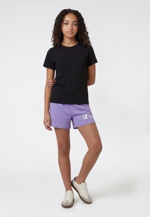 Teenager-Mädchen mit lockigem Haar, das ein schwarzes T-Shirt mit kurzen Ärmeln, lavendelfarbene Shorts mit Logo und weiße Turnschuhe trägt und vor einem schlichten Hintergrund steht.