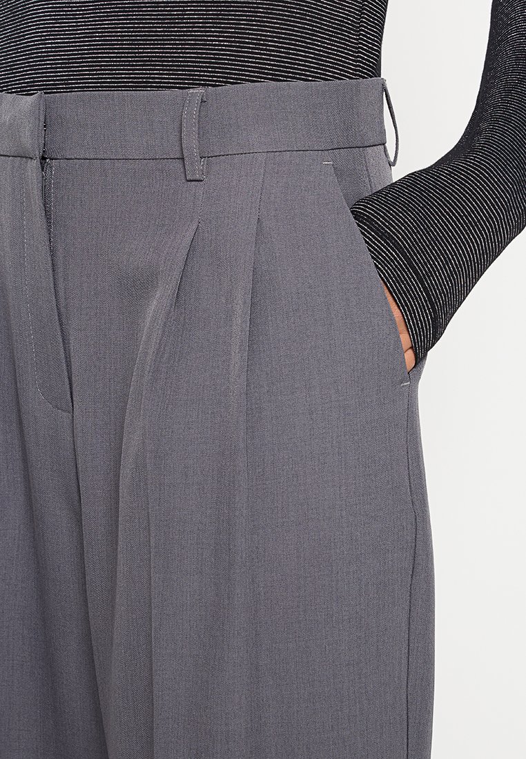 Graue, gefaltete Hose aus glattem Stoff mit tailliertem Schnitt, die über Seitentaschen und einen flachen Bund verfügt. Gestreiftes Oberteil ist teilweise sichtbar.