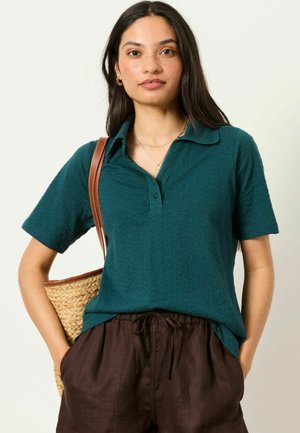 Woman with long dark hair wears teal short-sleeve polo, brown drawstring pants, gold jewelry, and carries straw tote over shoulder.