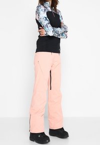 Giacca nera con fiori e zip, maniche lunghe, abbinata a pantaloni da neve rosa chiaro con cerniere, indossata con stivali da neve neri.