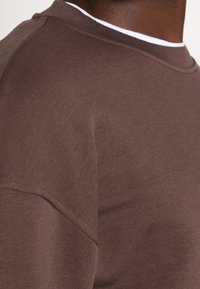 Sudadera de cuello redondo marrón hecha de una tela suave. Presenta una textura acanalada y un escote en contraste en blanco. Diseño simple y ajustado.