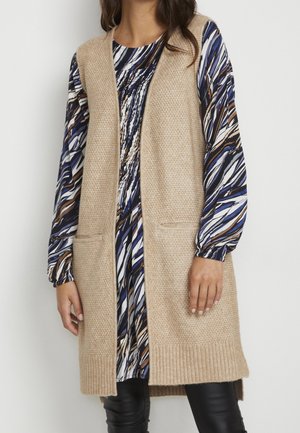 Femme portant un long gilet beige en maille par-dessus une robe à manches longues à motifs avec des rayures abstraites noires, blanches, bleues et marron.