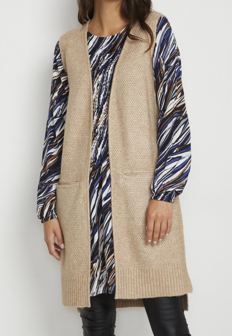 Femme portant un long gilet beige en maille par-dessus une robe à manches longues à motifs avec des rayures abstraites noires, blanches, bleues et marron.