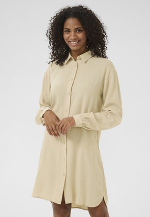 Button-up shirt dress in light beige fabric. Long sleeves with gathered cuffs, side slits at the hem, and a collared neckline.