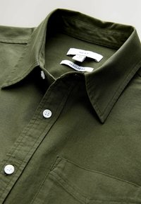 Cămașă din bumbac verde închis, cu guler care se button-down, nasturi albi, o buzunar și etichete care indică marca și stilul de potrivire. Design cu croială regular.
