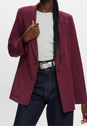 Femme portant un blazer bordeaux, un haut gris clair, un jean bleu foncé avec des surpiqûres orange et une boucle de ceinture argentée, avec les cheveux tressés sur l'épaule.