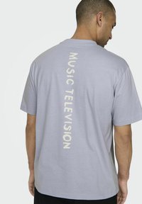 Lys grå t-shirt lavet af bomuld, med teksten "MUSIC TELEVISION" trykt lodret i en kontrasterende kremfarve på ryggen.