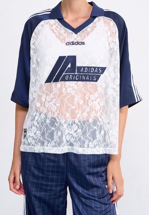 Osoba ubrana w granatowy dres Adidas z białą, kwiatową koronkową bluzką Adidas Originals na wierzchu.