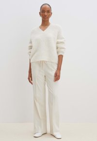 Cremefarbener, flauschiger V-Ausschnitt-Pullover mit gerippten Bündchen, kombiniert mit hoch taillierten, weiten weißen Hosen und glatten weißen Loafern.