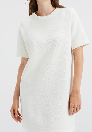 Mujer con un vestido de punto texturizado de manga corta blanca, mostrado desde los hombros hasta la mitad del muslo sobre un fondo liso claro.