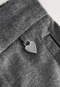 Nahaufnahme eines grauen Stoffes mit einer herzförmigen Metallklammer, auf der "Clarks" eingraviert ist und die an einer Schlaufe des Kleidungsstücks befestigt ist.