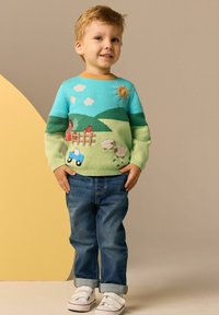 Pletený sveter s modro-zeleným dizajnom krajiny, ktorý zobrazuje slnko, oblaky a traktor, skombinovaný s modrými džínami a bielymi topánkami.