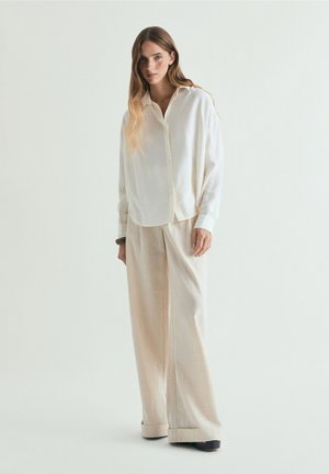 Junge Frau mit langen Haaren, die ein übergroßes weißes Hemd mit Knöpfen und weite beige Hosen trägt, steht vor hellem einfarbigem Hintergrund.