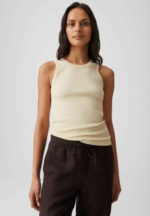 Femme aux longs cheveux foncés portant un débardeur beige côtelé sans manches et un pantalon marron foncé à cordon, debout devant un fond clair uni.