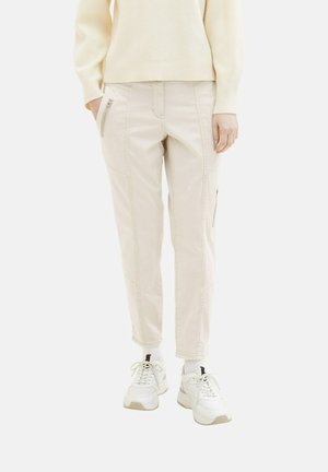 Personne portant un pantalon beige clair avec des poches zippées, des chaussettes blanches, des baskets blanches et un pull côtelé de couleur crème.