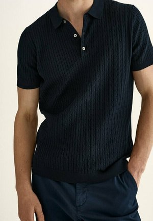 Man wearing a dark navy short-sleeve cable-knit polo shirt with buttons, paired with dark navy pants, one hand in pocket.
