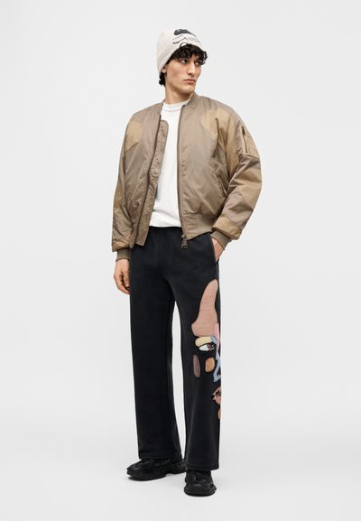 Μπεζ μπουφάν bomber με γυαλιστερό φινίρισμα, σε συνδυασμό με μαύρο παντελόνι φόρμας με πολύχρωμη αφηρημένη κεντητική. Μαύρα αθλητικά παπούτσια συμπληρώνουν την εμφάνιση.