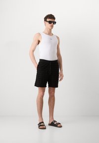 Witte tanktop, zwarte textuur shorts met horizontale strepen, zwarte zonnebril en donkere sandalen. Simpele, functionele strandoutfit.