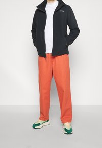 Giacca in pile nera con zip e due tasche, abbinata a pantaloni arancioni larghi. La calzatura presenta accenti verde e crema.