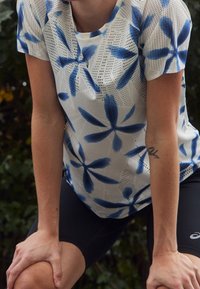 Graphic athletic top featuring white mesh fabric with blue floral patterns. Short sleeves and round neckline. Paired with black shorts.