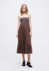 Γυναίκα που στέκεται φορόντας ένα καφέ μίντι φόρεμα χωρίς τιράντες με σμιλευμένο μπλε και καφέ μπούστο και μαύρα ψηλοτάκουνα mule, σε λευκό φόντο.