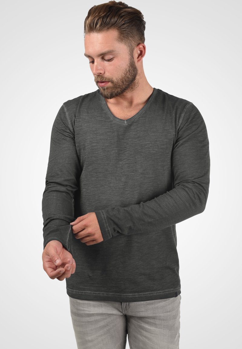 Cămașă lungă gri cu mâneci lungi și decolteu în V, având un material texturat și mâneci strâmte. Modelul își ajustează lungimea mânecii.
