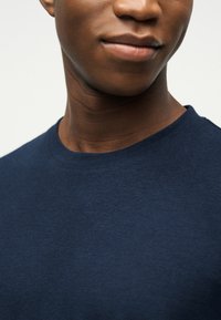 Νεαρό άτομο με σκουρόχρωμο δέρμα που φοράει απλό μπλε σκούρο T-shirt με στρογγυλή λαιμόκοψη, χαμογελώντας ελαφρώς, μπροστά σε ανοιχτό φόντο.