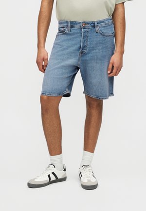 Mężczyzna w jasnoniebieskich dżinsowych szortach, białych skarpetkach i białych trampkach z czarnymi paskami, stojący na jednolitym tle.