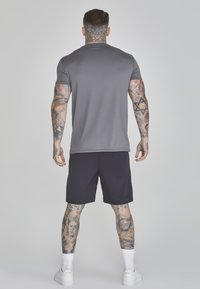 Cămașă gri cu mânecă scurtă și pantaloni scurți negri. Tatuaje vizibile pe brațe și picioare. Adidași albi și șosete scurte completează ținuta.