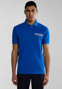 Camisa polo azul de algodón con cuello clásico, mangas cortas y un logo impreso en el lado izquierdo del pecho. Color sólido, textura suave.