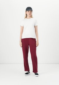 Giovane che indossa una maglietta bianca, pantaloni bordeaux, cappellino nero e sneakers bianco e nere, in piedi contro uno sfondo bianco uniforme.