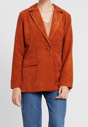 Blazer en velours côtelé orange rouille avec deux poches avant à rabat porté sur un top beige et un jean bleu, visible du cou aux hanches.