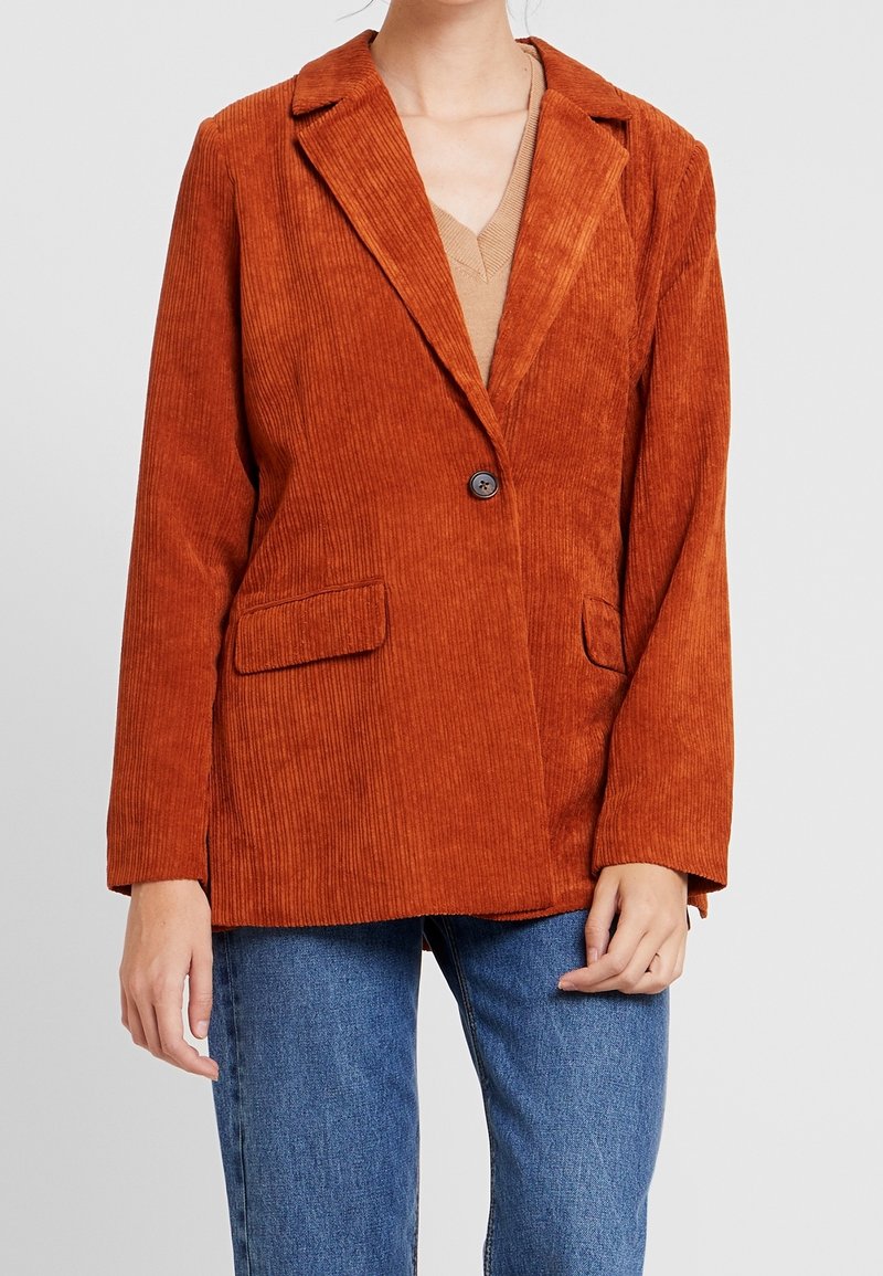 Blazer en velours côtelé orange rouille avec deux poches avant à rabat porté sur un top beige et un jean bleu, visible du cou aux hanches.