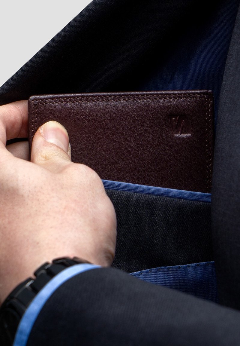 Brown leather wallet is partially pulled from a dark jacket pocket. The wallet features a subtle embossed logo and neat stitching along the edges.
