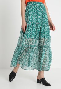 Jupe midi à volants en vert, avec un motif floral. Fabriquée en tissu léger et transparent ; taille élastique ; associée à des mules noires.