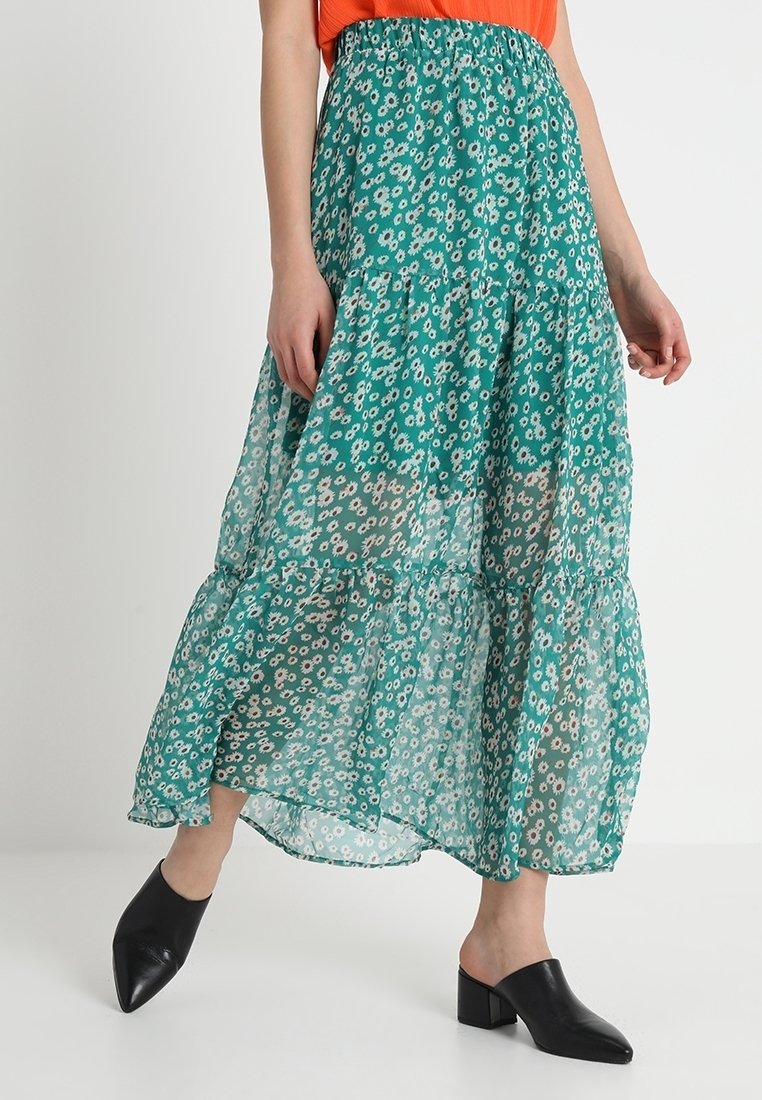 Jupe midi à volants en vert, avec un motif floral. Fabriquée en tissu léger et transparent ; taille élastique ; associée à des mules noires.
