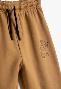 Pantalones de chándal beige con un acabado texturizado, que presentan cordones negros y un logo NY impreso con un león. Cintura elástica para mayor comodidad.